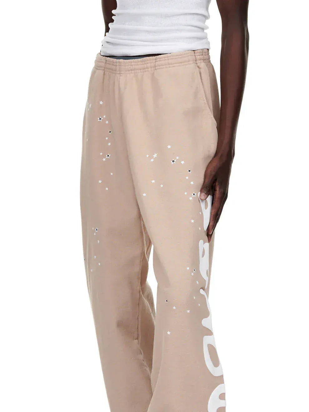 A person wearing 100% Cotton Sp5der Sweatpants for Men—beige streetwear jogger pants with side logo graphic and a relaxed fit—paired with a white ribbed tank top.