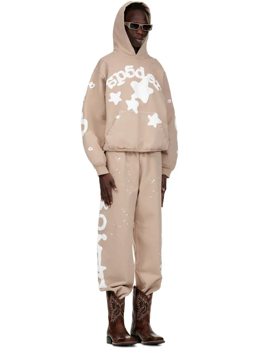 A person in 100% Cotton Sp5der Sweatpants for Men—beige streetwear jogger pants with side logo graphic—plus a beige hoodie, dark sunglasses, and brown cowboy boots, stands against a plain background.