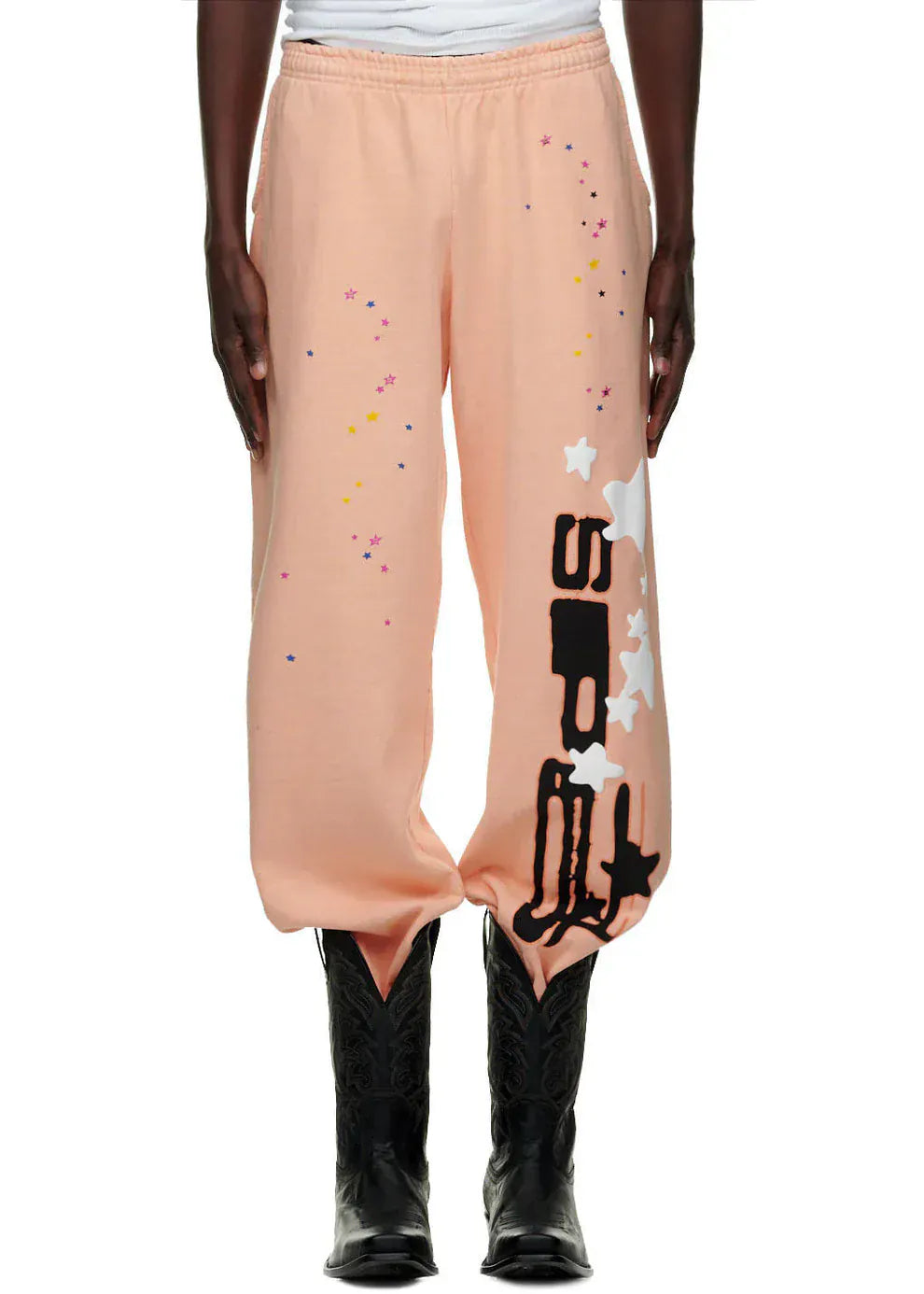 Person wearing 100% cotton Sp5der unisex peach jogger sweatpants with vertical logo and star graphics, relaxed fit, styled with black cowboy boots—a standout streetwear look.