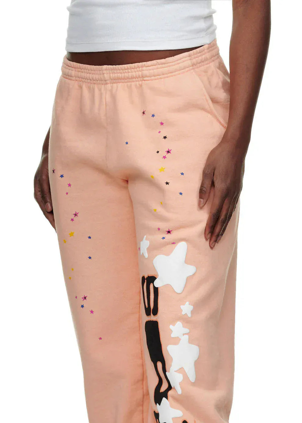 Model wears 100% Cotton Sp5der Sweatpants—Unisex Peach Jogger Pants with Vertical Logo and Star Graphics, relaxed fit—paired with a white crop top. Only lower torso and one arm are visible, ideal for a bold streetwear statement.