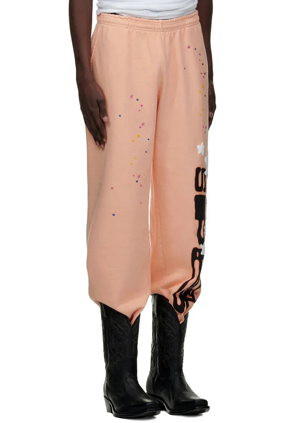 Wearing 100% Cotton Sp5der Sweatpants—Unisex Peach Jogger Pants with a vertical logo and star graphics, relaxed fit—a person pairs these streetwear pants with black cowboy boots. Only their lower body is visible.
