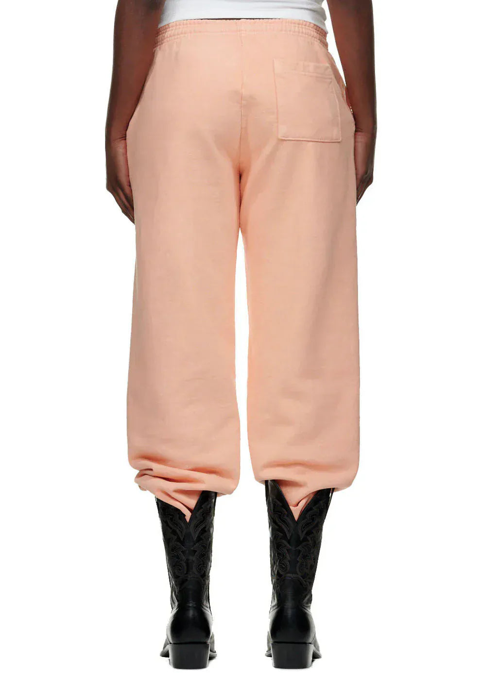 Model wearing relaxed-fit 100% cotton Sp5der sweatpants—unisex peach jogger pants with vertical logo and star graphics—styled with black cowboy boots, photographed from the back against a plain white background.