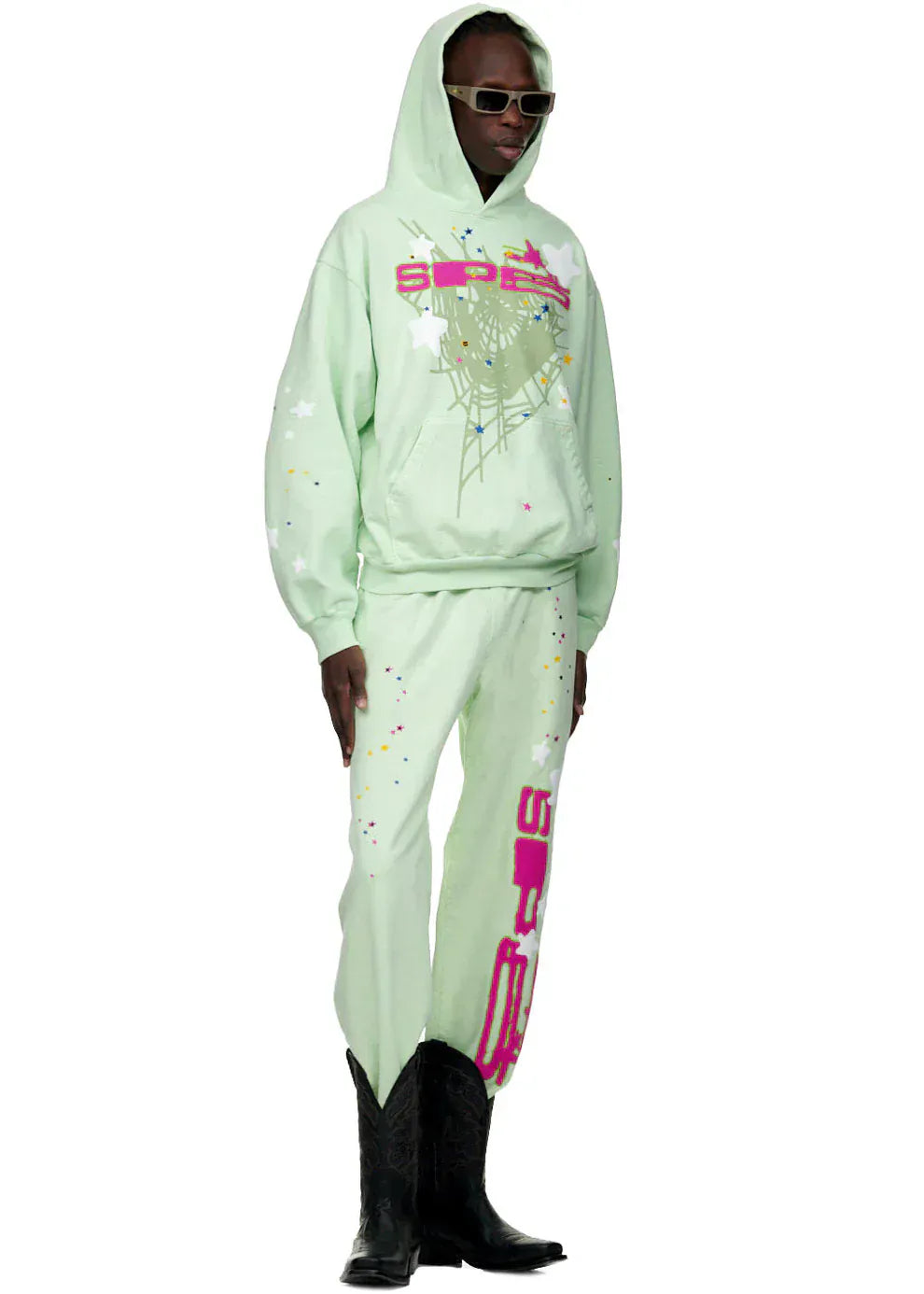 A person wears 100% Cotton Sp5der Sweatpants – Unisex Mint Green Jogger Pants with vertical logo and star graphics, black cowboy boots, and sunglasses, highlighting bold streetwear style against a white background.