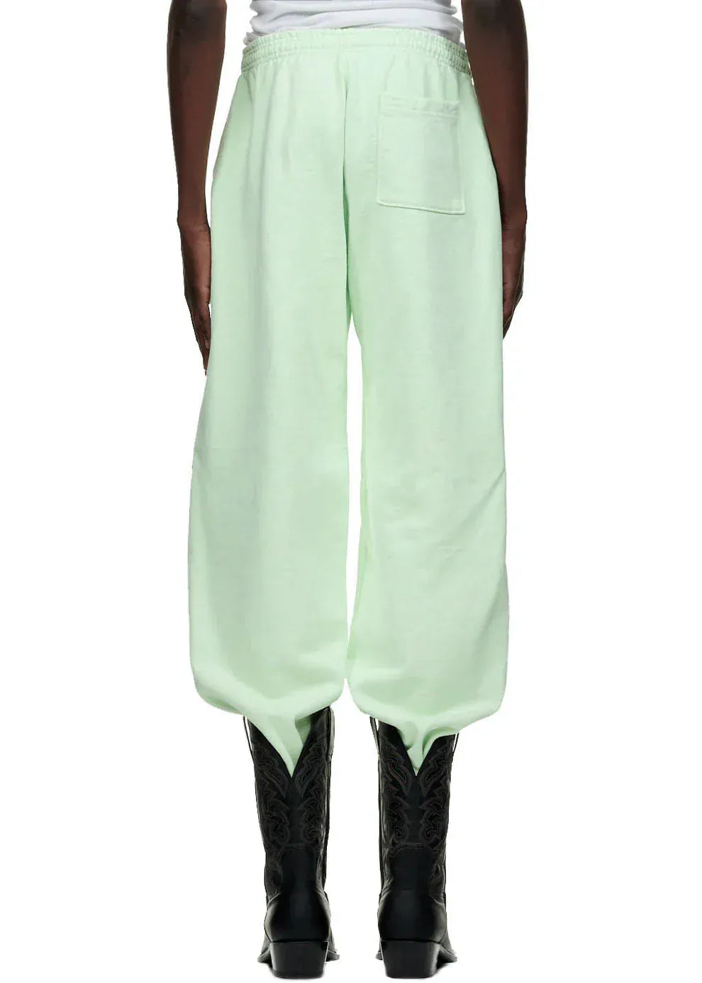 A person sports unisex Sp5der mint green 100% cotton jogger pants with vertical logo and star graphics, tucked into black cowboy boots—an effortlessly cool streetwear style shown from the back.