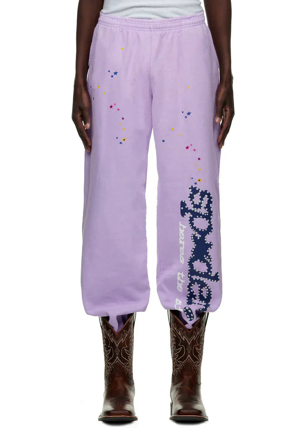 Someone wearing 100% cotton Sp5der unisex lavender jogger pants with vertical logo and star details, styled with brown cowboy boots for a unique streetwear look.
