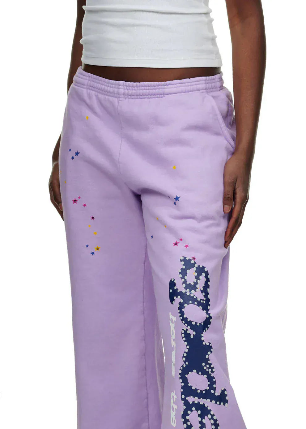 A person wearing a white tank top and 100% Cotton Sp5der Unisex Lavender Jogger Pants featuring a vertical logo and star details, shown from mid-torso to knees.