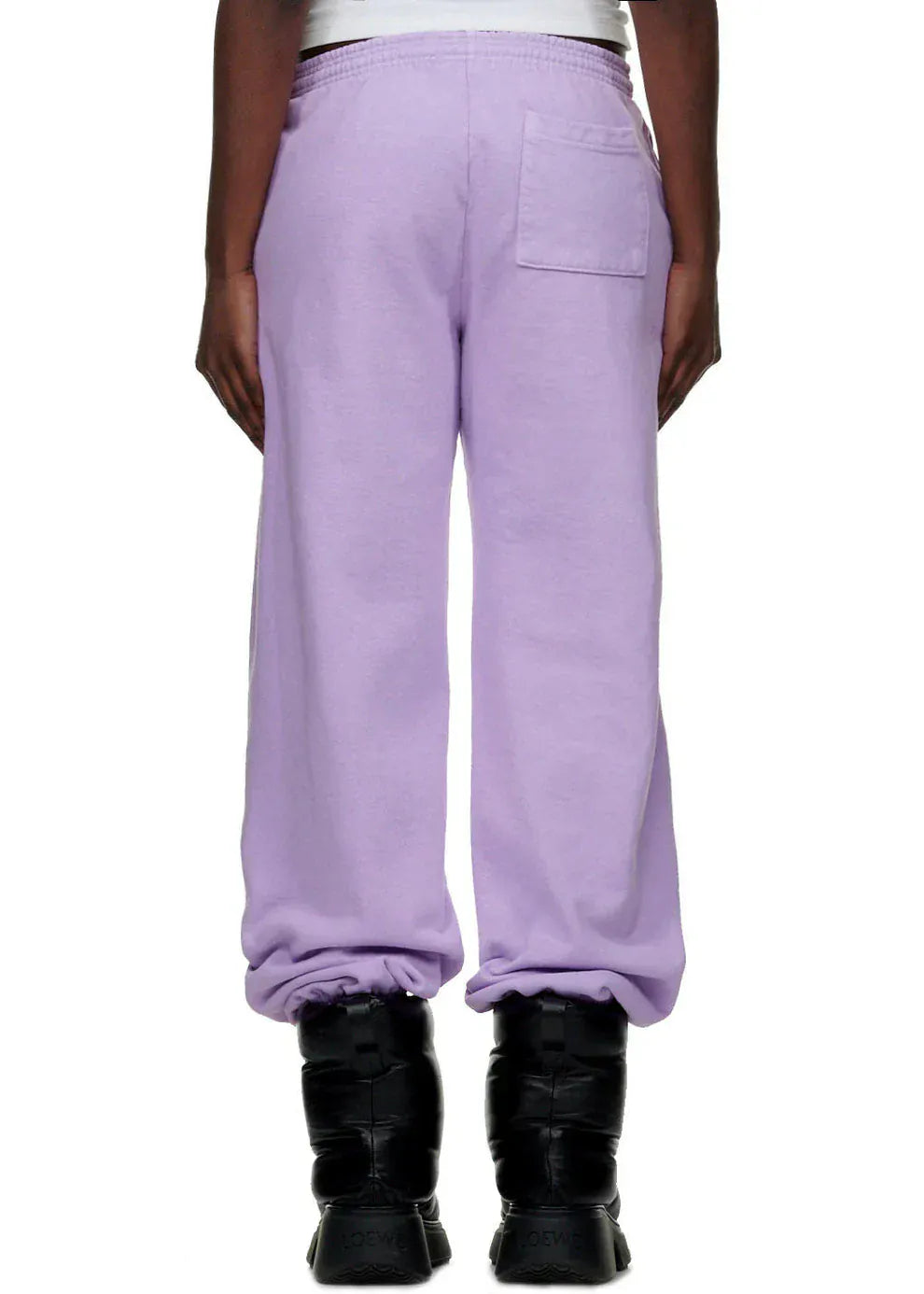 A person wearing 100% cotton Sp5der unisex lavender jogger pants with a vertical logo and star details, shown from the back with black boots. The relaxed-fit streetwear features an elastic waistband and a single back pocket.