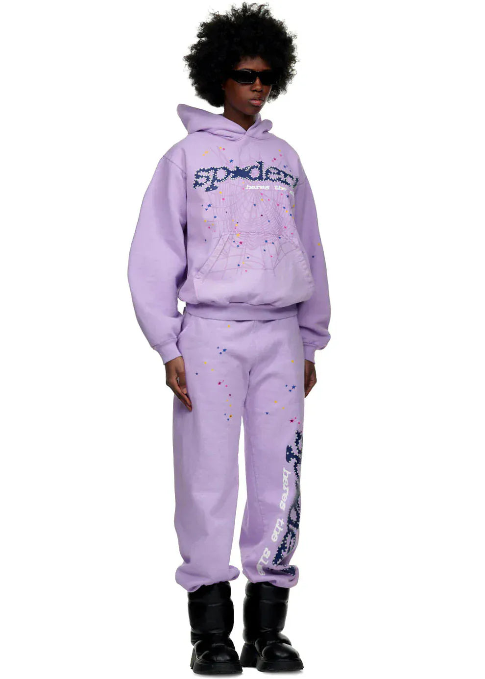 A person showcases unisex streetwear in a lavender hoodie, 100% Cotton Sp5der Sweatpants—Unisex Lavender Jogger Pants with vertical logo and star details, relaxed fit—black boots, and sunglasses against a white background.