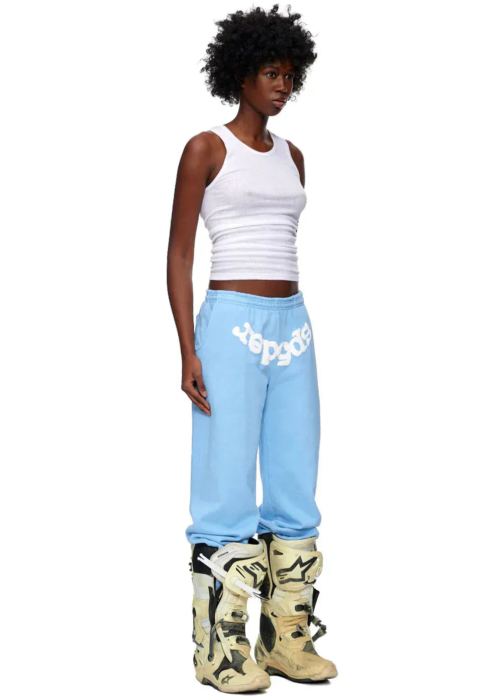 A person in a white ribbed tank top, Sp5der Unisex Light Blue Cotton Sweatpants with front logo print, and beige motocross boots stands against a plain white background, highlighting relaxed streetwear style.