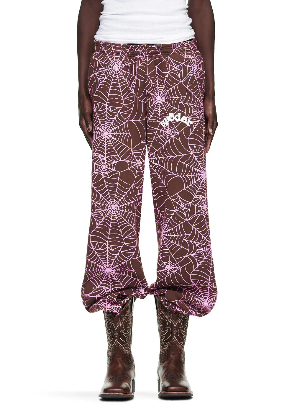 A person wears 100% Cotton Sp5der Sweatpants—Unisex Brown Jogger Pants with all-over pink spider web print and a visible white logo—paired with a white shirt and brown cowboy boots.