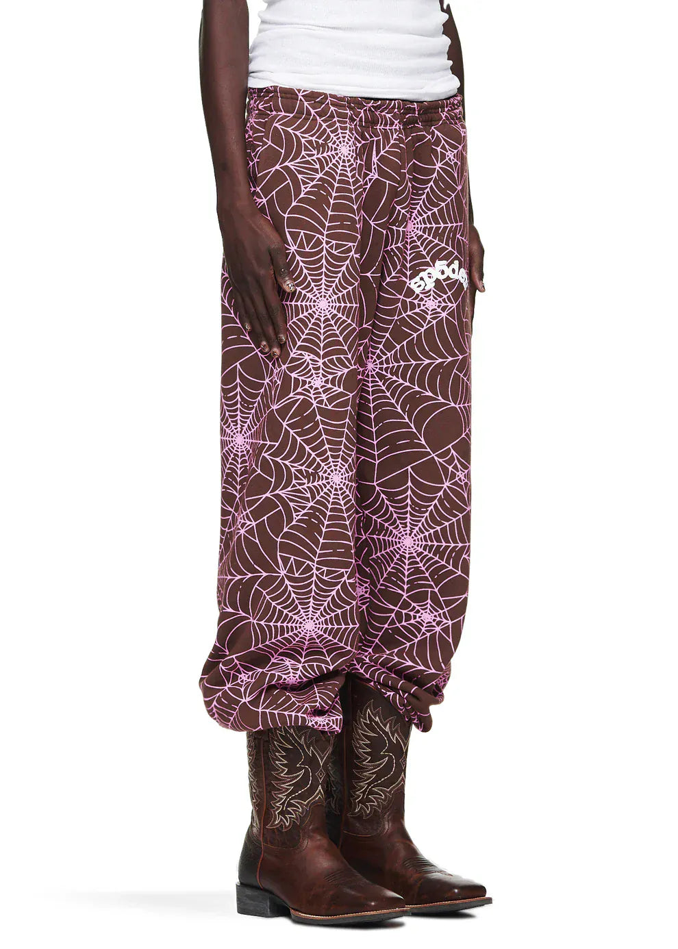 A person wearing 100% Cotton Sp5der Sweatpants – Unisex Brown Jogger Pants with All-Over Spider Web Print, a white tank top, and brown cowboy boots.