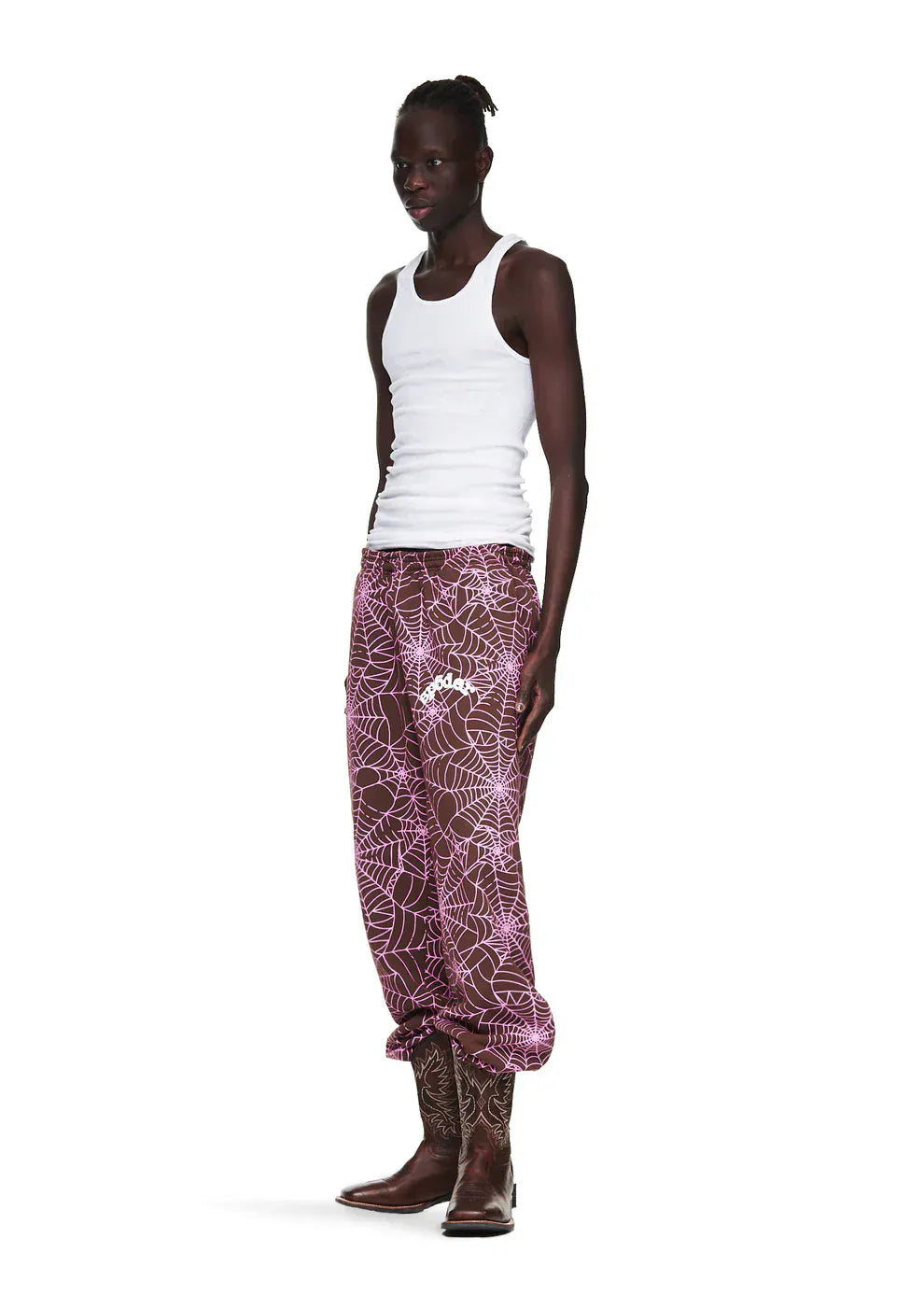 A person stands against a plain white background wearing a white tank top, brown cowboy boots, and 100% Cotton Sp5der Sweatpants—unisex brown jogger pants featuring an all-over spider web print and relaxed fit.