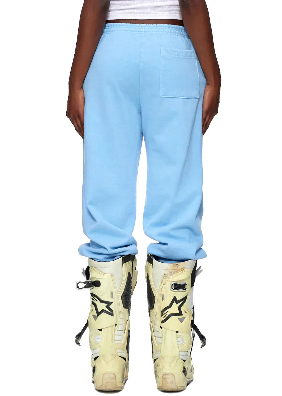 A person wearing Sp5der Sweatpants – Unisex Light Blue Cotton Sweatpants with Front Logo Print and white motocross boots stands facing away from the camera, highlighting bold streetwear style.