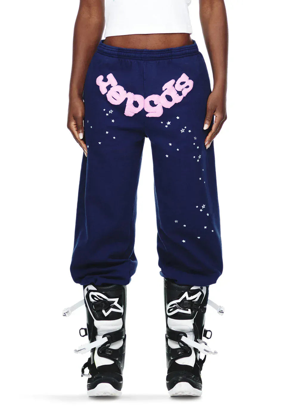 A person wears Sp5der 100% cotton unisex navy blue jogger pants with a pink logo and star graphics, paired with black and white motocross boots. Only the lower torso and legs are shown, highlighting the relaxed streetwear style.