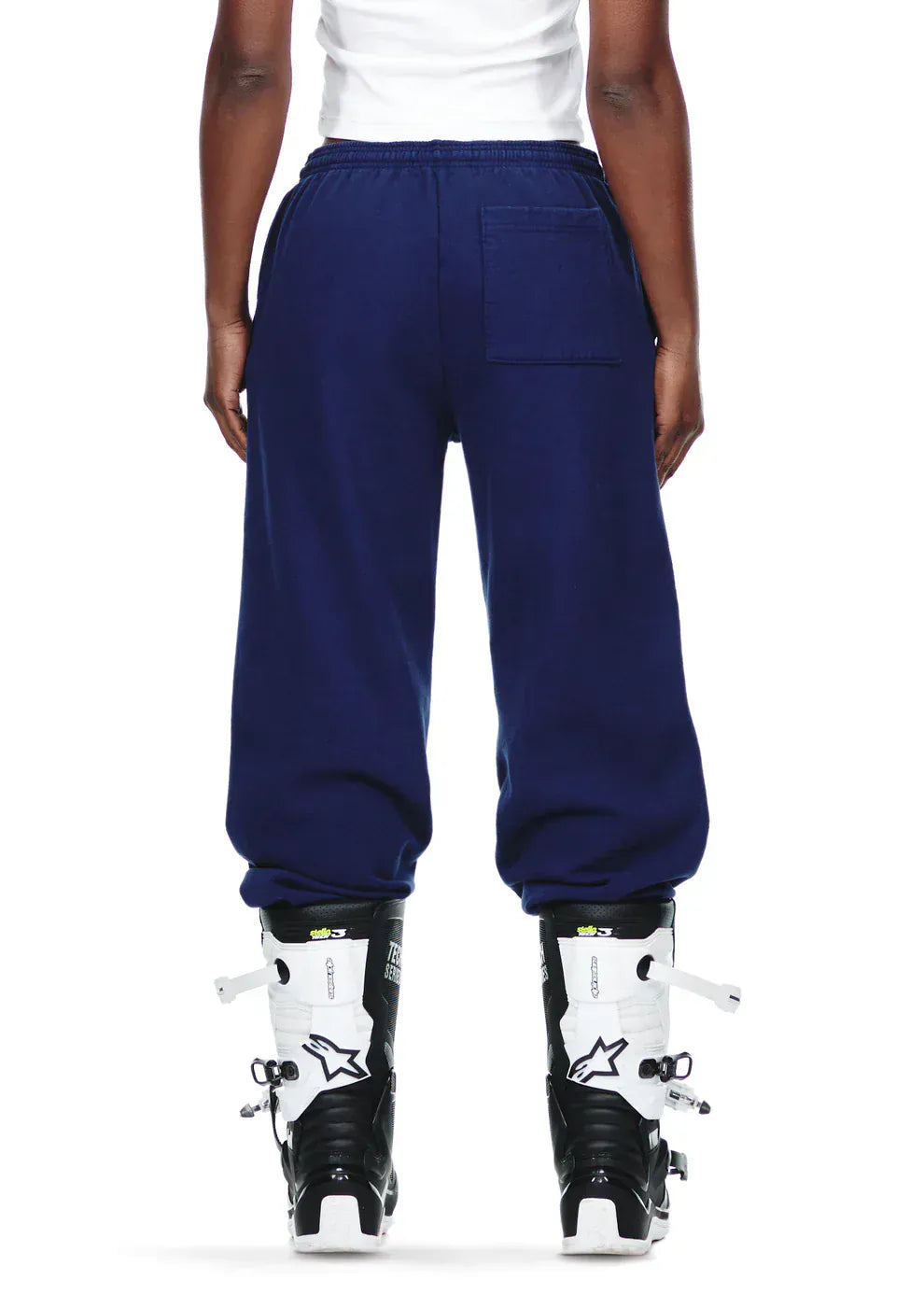 Person standing with their back to the camera, wearing a white shirt and 100% Cotton Sp5der Sweatpants—Unisex Navy Blue Jogger Pants with Pink Logo and Star Graphics, Relaxed Fit—paired with black and white ski boots.
