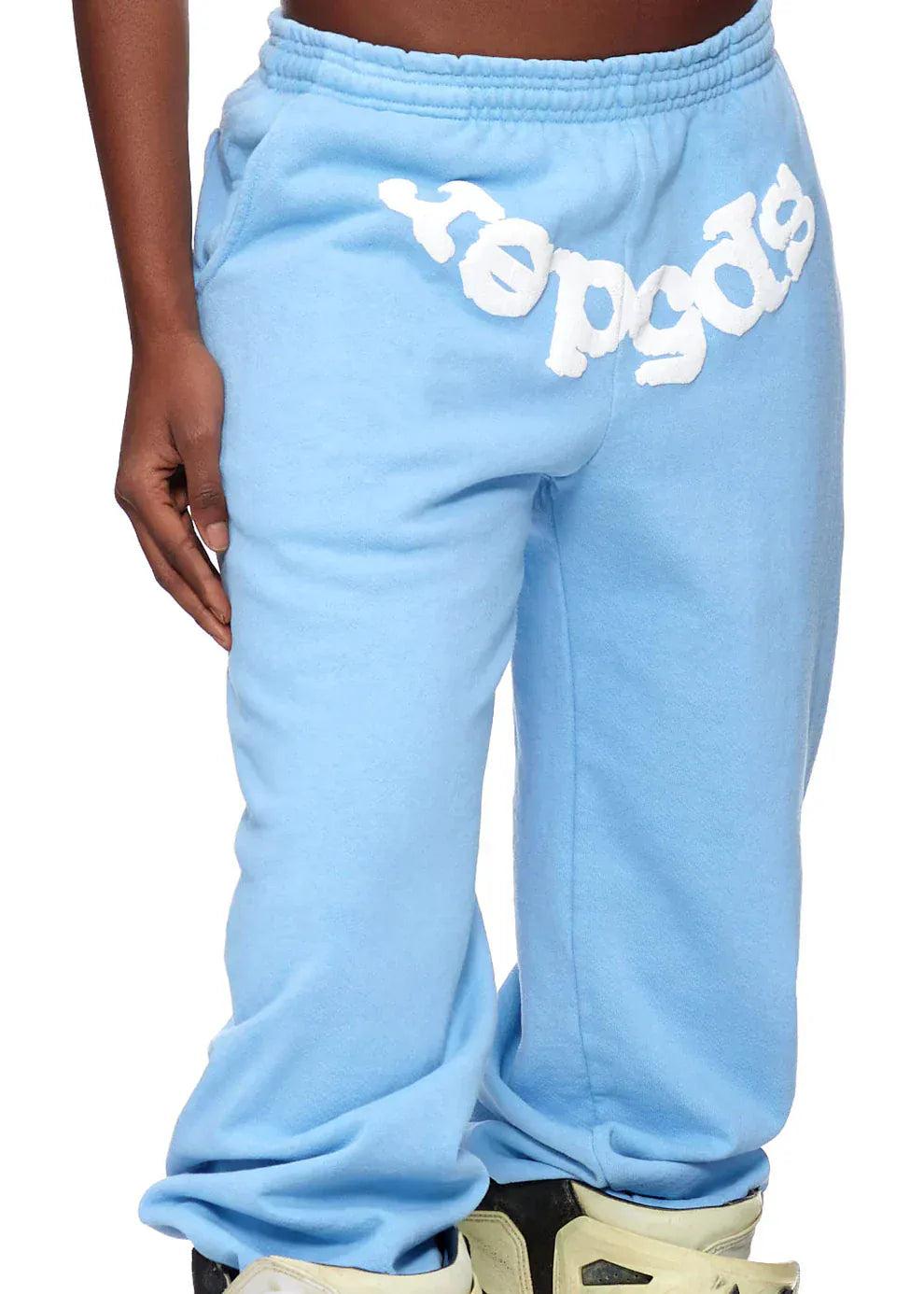 A person in unisex light blue Sp5der Sweatpants with front logo print, styled with light sneakers—a fresh streetwear look.