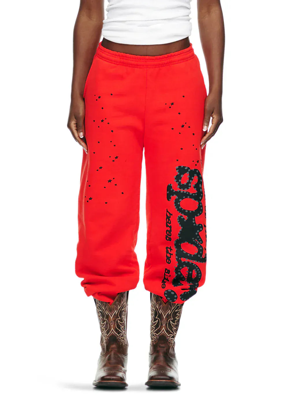 A person wears 100% Cotton Sp5der Sweatpants—Unisex Red Jogger Pants with Black Logo and Star Graphics, a white shirt, and brown cowboy boots against a plain background for a casual streetwear look.