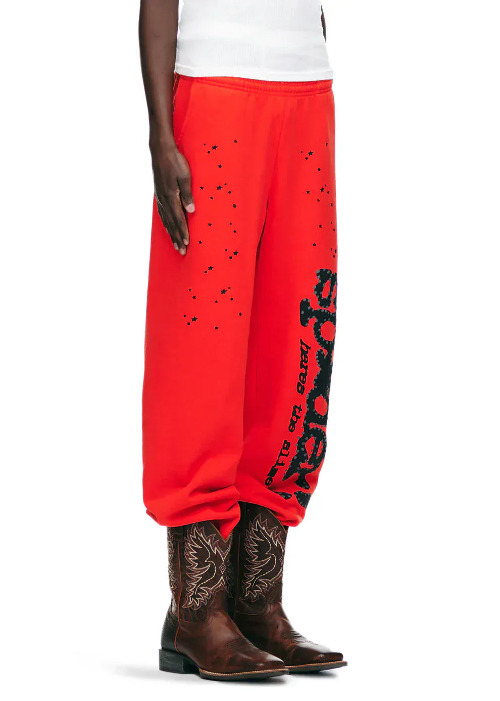 Wearing 100% Cotton Sp5der Sweatpants—Unisex Red Jogger Pants with Black Logo and Star Graphics, Relaxed Fit—paired with brown cowboy boots and a white top creates a bold streetwear statement.