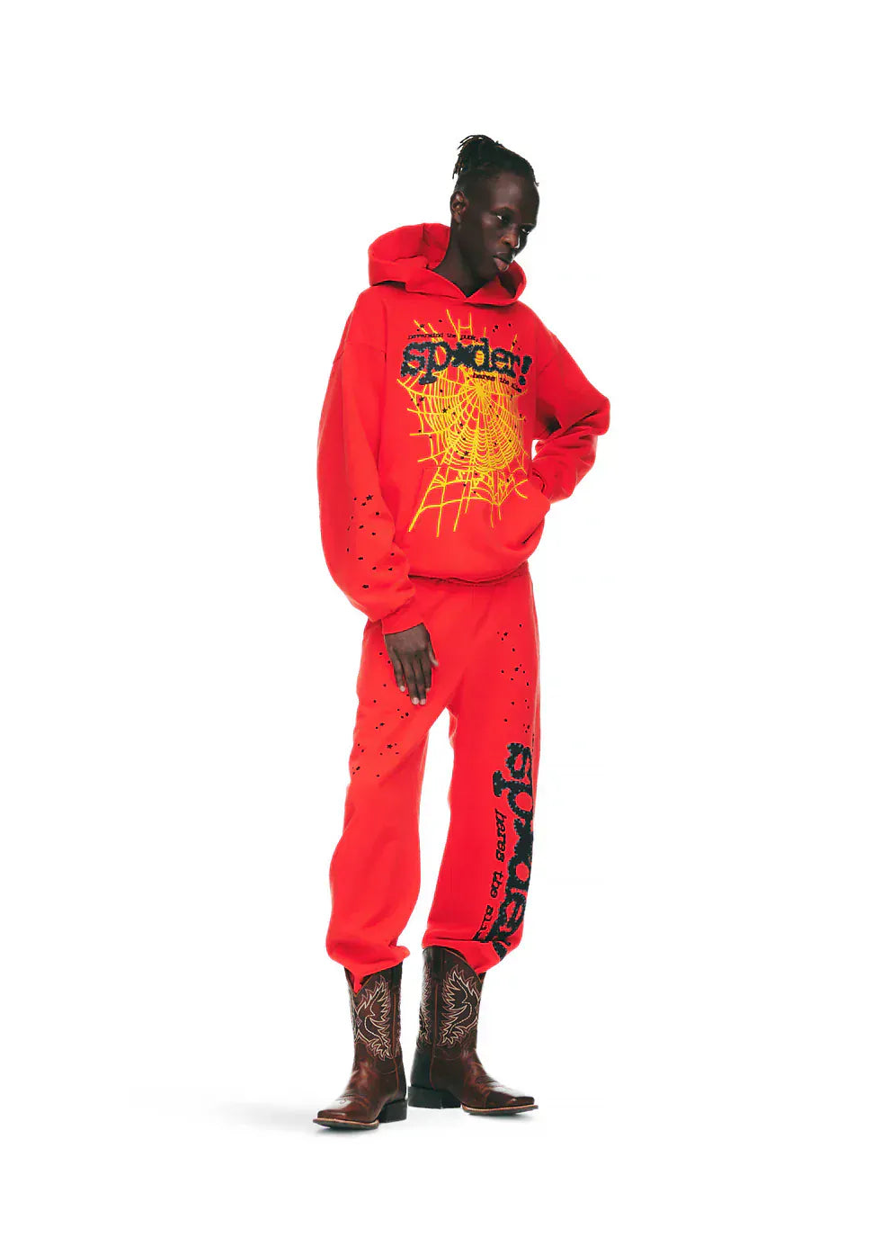 Person wearing a bright red hoodie and 100% Cotton Sp5der Sweatpants—Unisex Red Jogger Pants with Black Logo and Star Graphics, Relaxed Fit—paired with brown cowboy boots, standing against a white background.