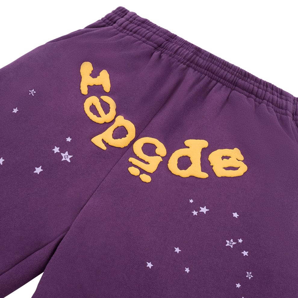 Close-up of 100% cotton Sp5der Sweatpants—unisex purple jogger pants with a yellow logo and scattered star graphics, featuring a relaxed fit for bold streetwear style.