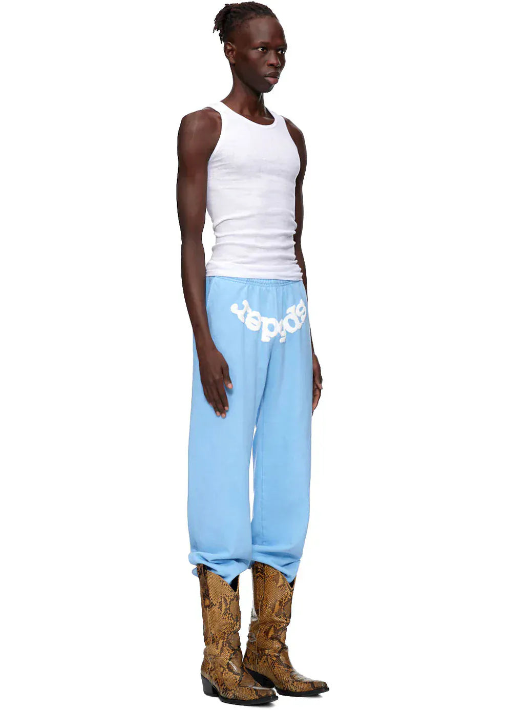 Person in profile wearing a white tank top, Sp5der Sweatpants—unisex light blue cotton sweatpants with a front logo print—and brown snake-print boots, creating a modern streetwear look against a plain background.
