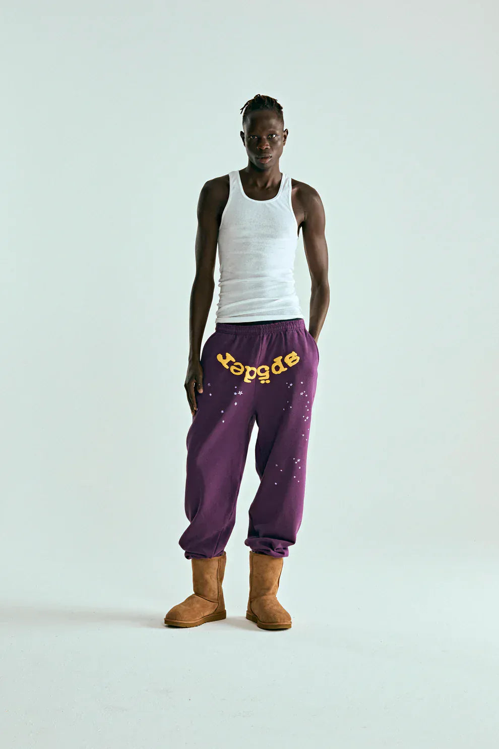 A person stands against a plain background in a white tank top, tan boots, and 100% cotton Sp5der Sweatpants—unisex purple jogger pants with yellow logo, star graphics, and a relaxed fit for an effortless streetwear vibe.