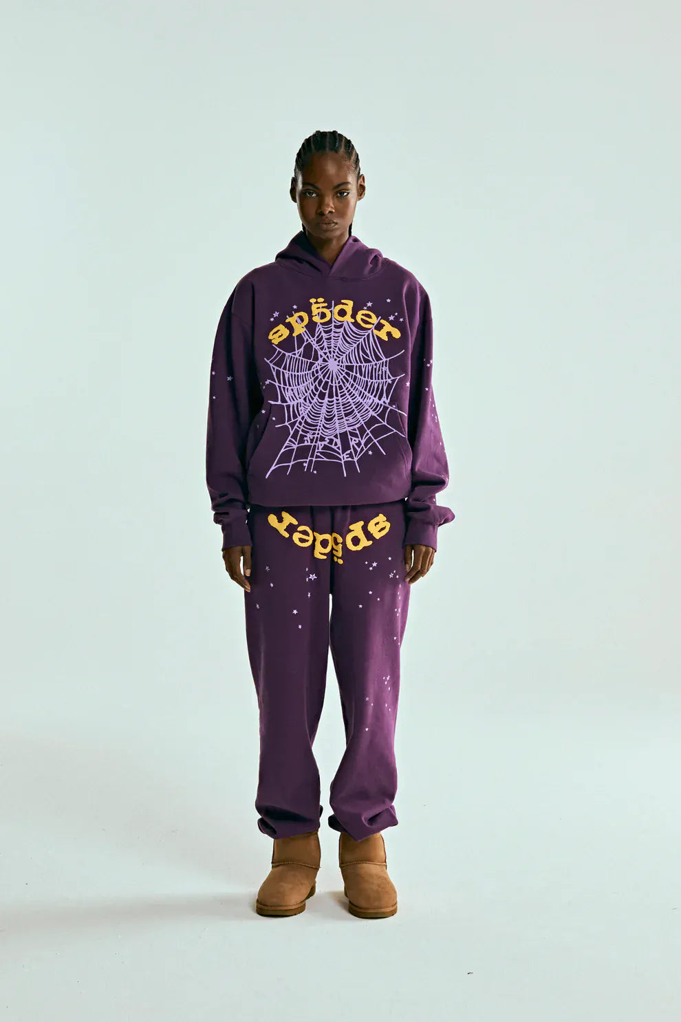 A person stands facing forward in a purple hoodie and 100% Cotton Sp5der Sweatpants—unisex purple jogger pants with yellow logo and star graphics—in a relaxed fit, paired with tan boots, against a plain light background.