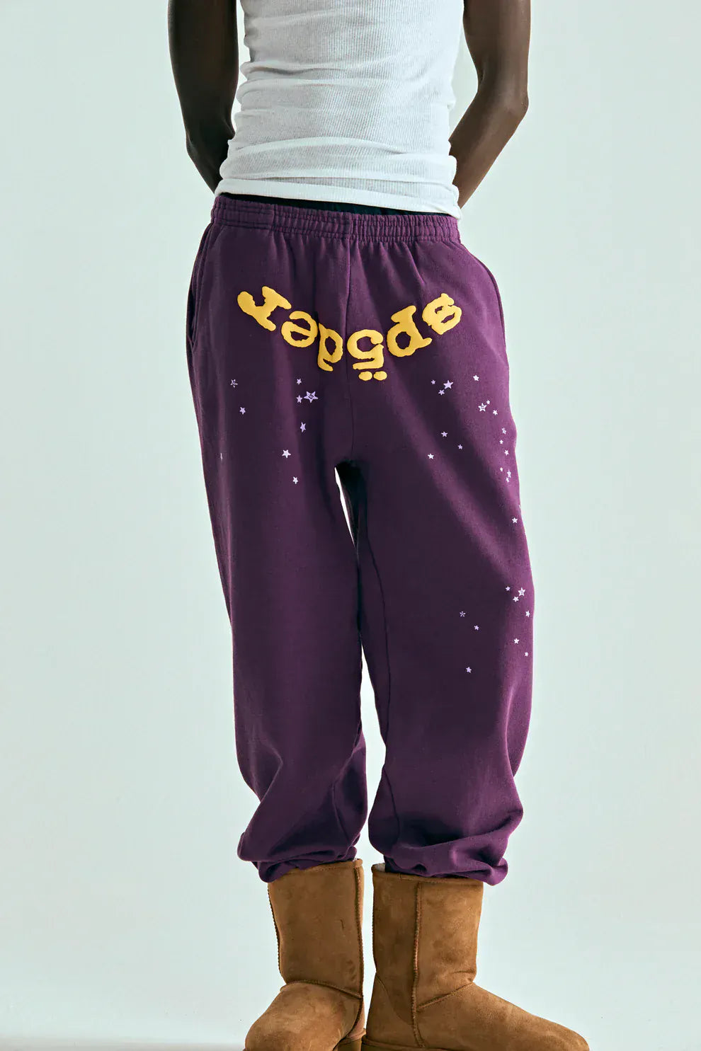 A person wears a white tank top, brown boots, and 100% Cotton Sp5der Sweatpants—Unisex Purple Jogger Pants with yellow logo and star graphics—showcasing relaxed-fit streetwear style against a plain background.