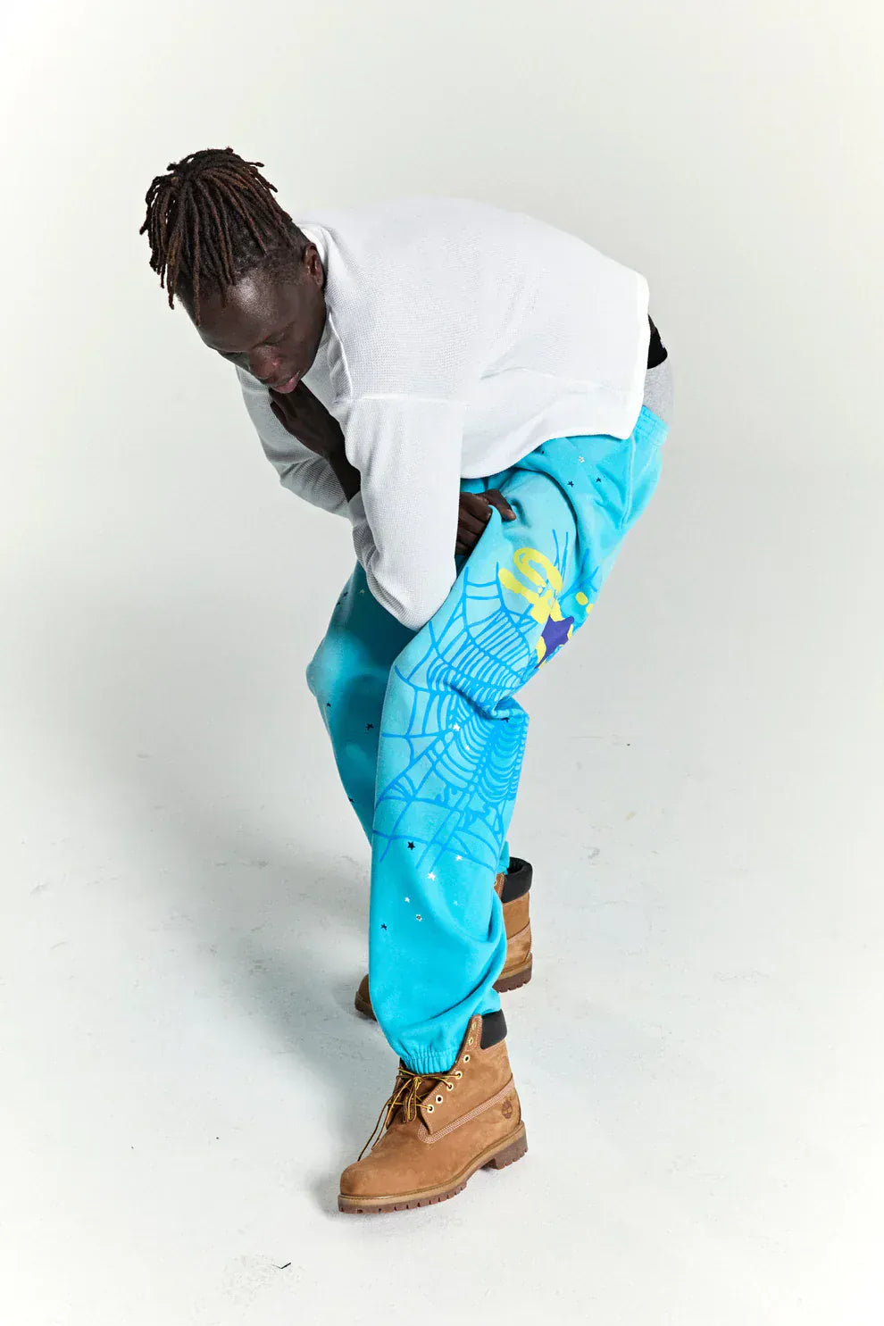A person bends forward against a plain white background, showcasing the 100% Cotton Sp5der Unisex Blue Jogger Pants with spider web graphic, star details, and a relaxed fit for casual streetwear style.