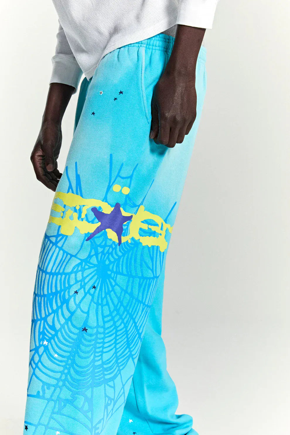 A person wearing 100% cotton Sp5der sweatpants—unisex blue jogger pants with spider web graphic, star details, and a relaxed fit—styled with a white long-sleeve top for the perfect streetwear look.