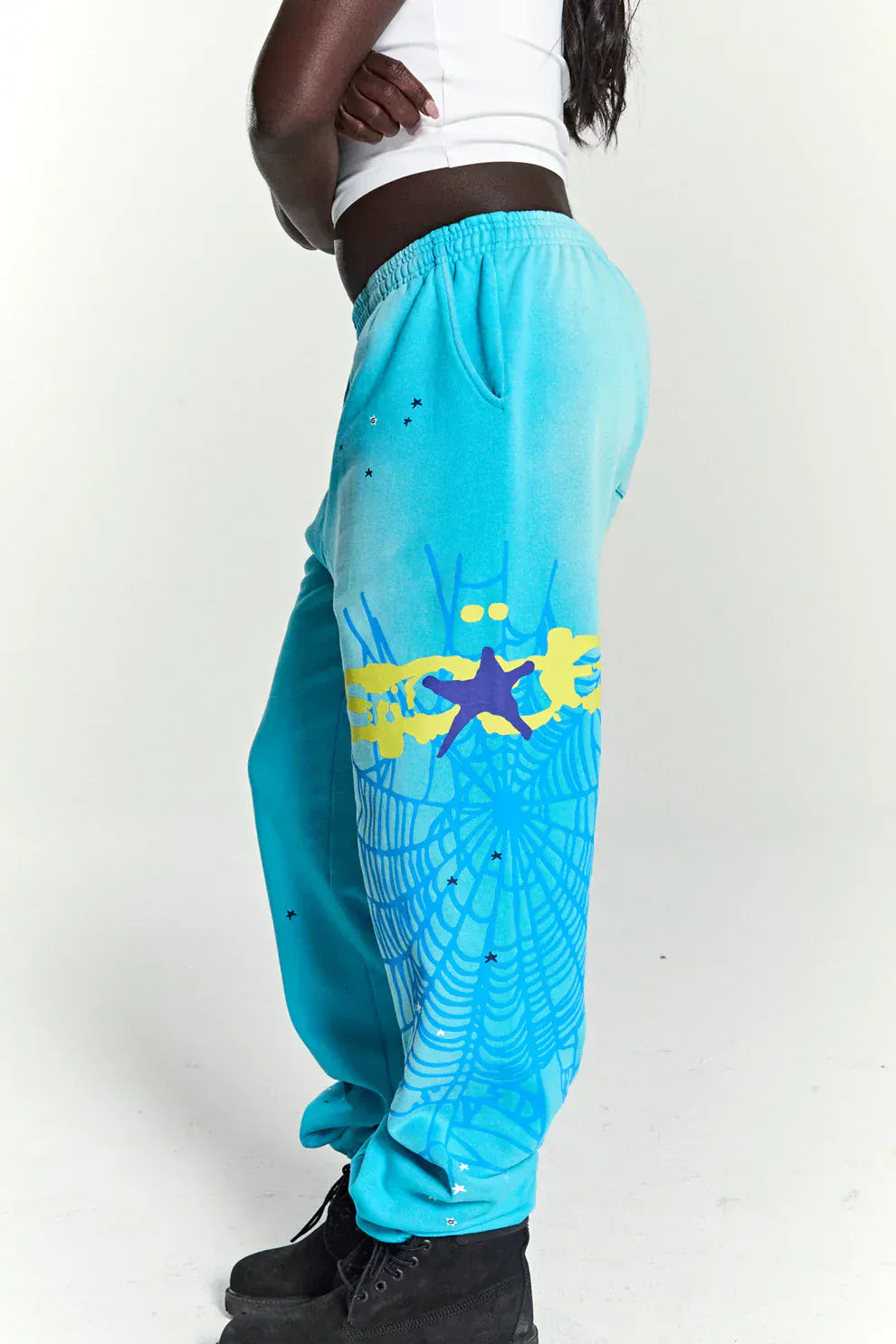 Person wearing 100% Cotton Sp5der Sweatpants—Unisex Blue Jogger Pants with spider web graphic and star details, relaxed fit—styled with a white crop top and black boots for cool streetwear against a white background.