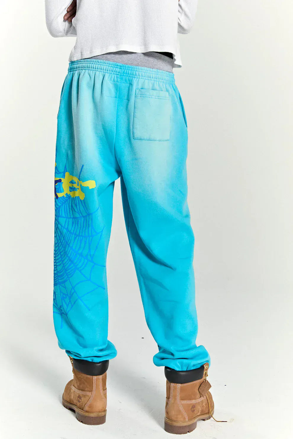 Person wears 100% Cotton Sp5der Unisex Blue Sweatpants with spider web graphics and star details, tan boots, and a white shirt—shown from the back against a plain background for a relaxed streetwear look.