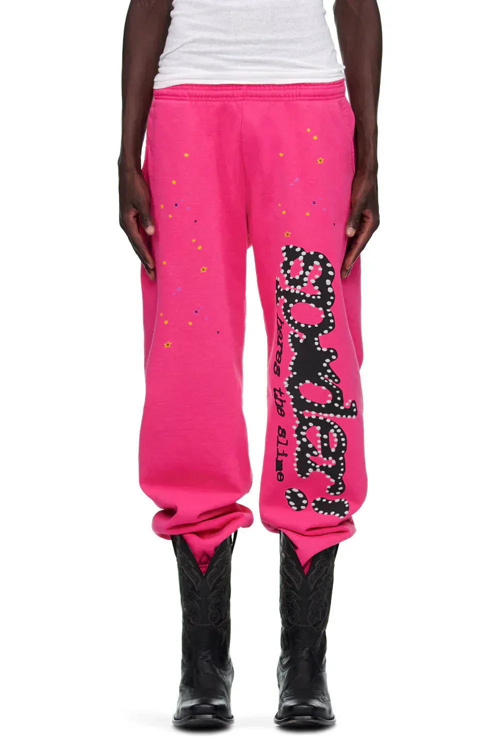 Someone stands against a plain background wearing Sp5der Sweatpants—unisex pink cotton wide leg sweatpants with a graphic logo print—paired with a white shirt and black cowboy boots.