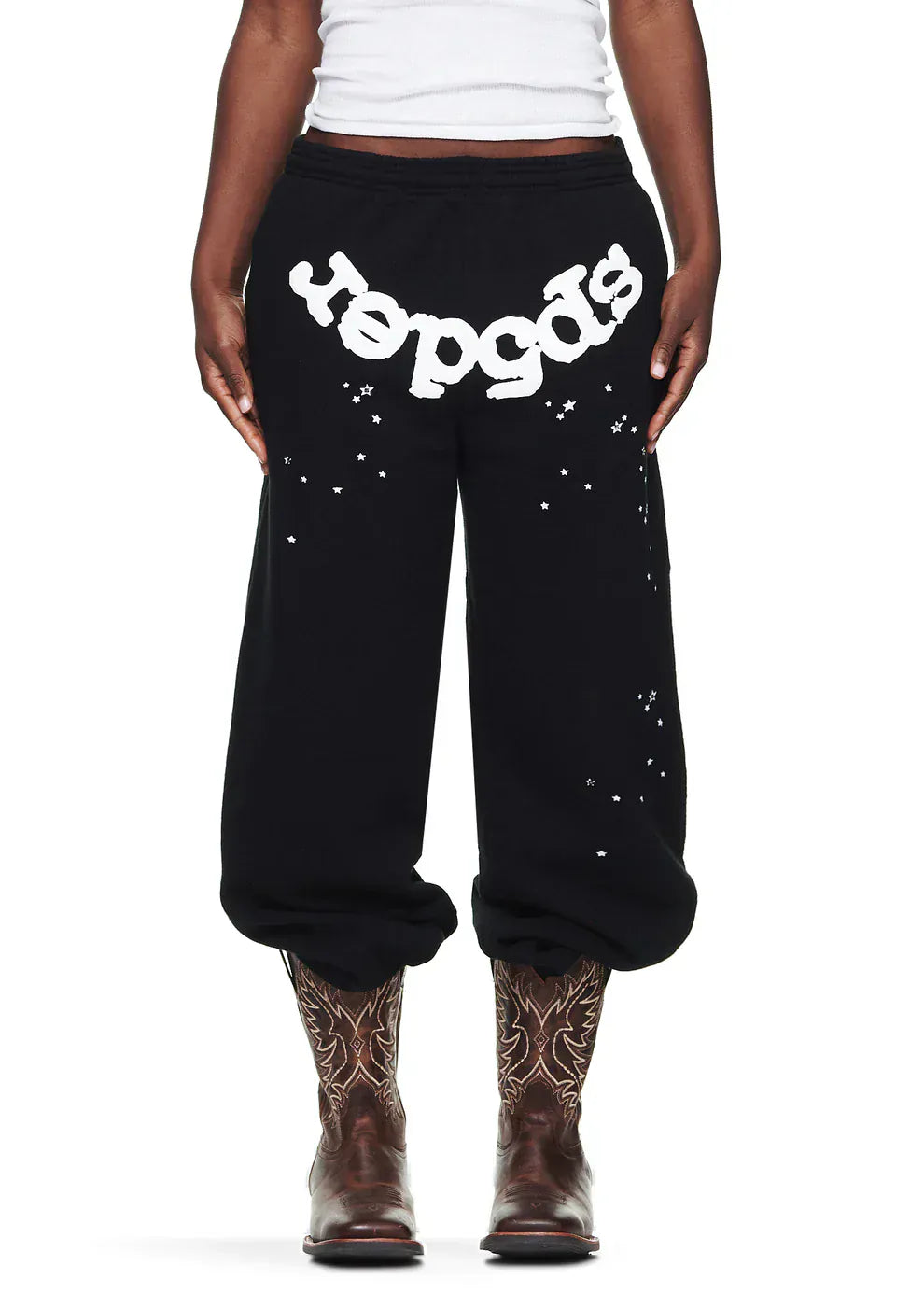 Wearing Sp5der Sweatpants—unisex black cotton with front logo and star print—a white tank top, and brown cowboy boots, the person stands against a plain background, combining edgy streetwear with Western flair.