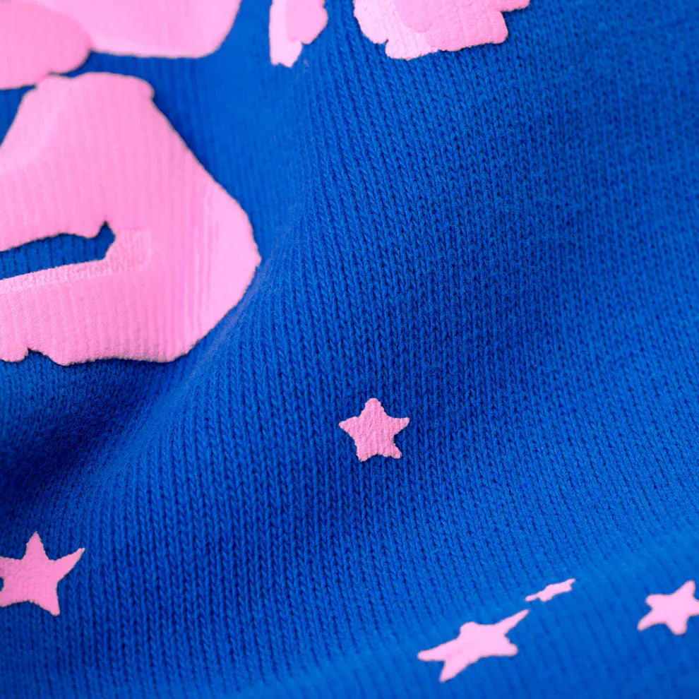 Close-up of bold pink logo and star accents on textured blue fabric, highlighting the design of 100% Cotton Sp5der Sweatpants—Unisex Blue Jogger Pants with a relaxed fit, ideal for streetwear enthusiasts and cotton jogger pants fans.