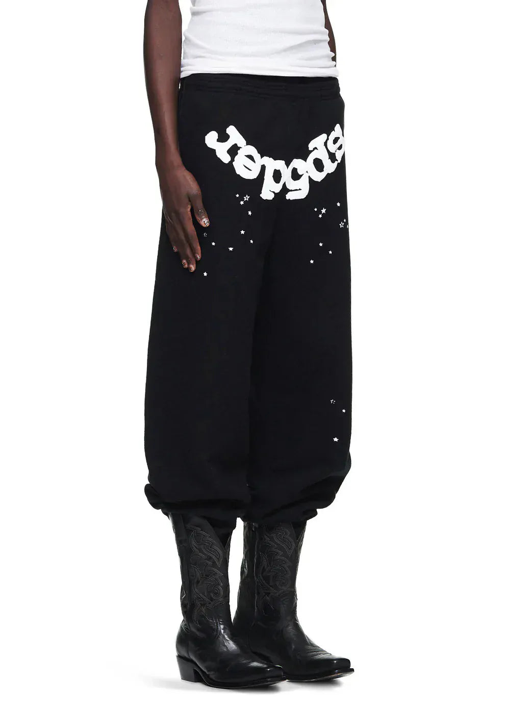 Wearing Sp5der Sweatpants—unisex black cotton with front logo and star print—paired with black cowboy boots and a white top, creates a bold streetwear look.