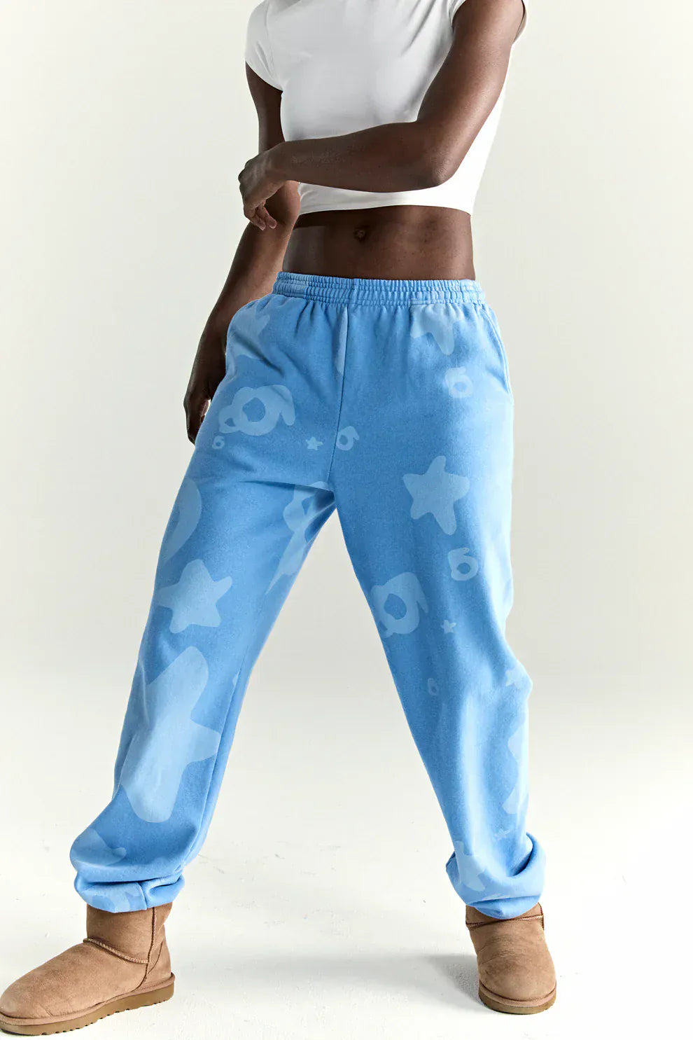 A person wearing a white cropped t-shirt, Sp5der Light Blue All-Over Print Sweatpants (100% cotton unisex streetwear joggers), and tan boots stands against a plain light background.