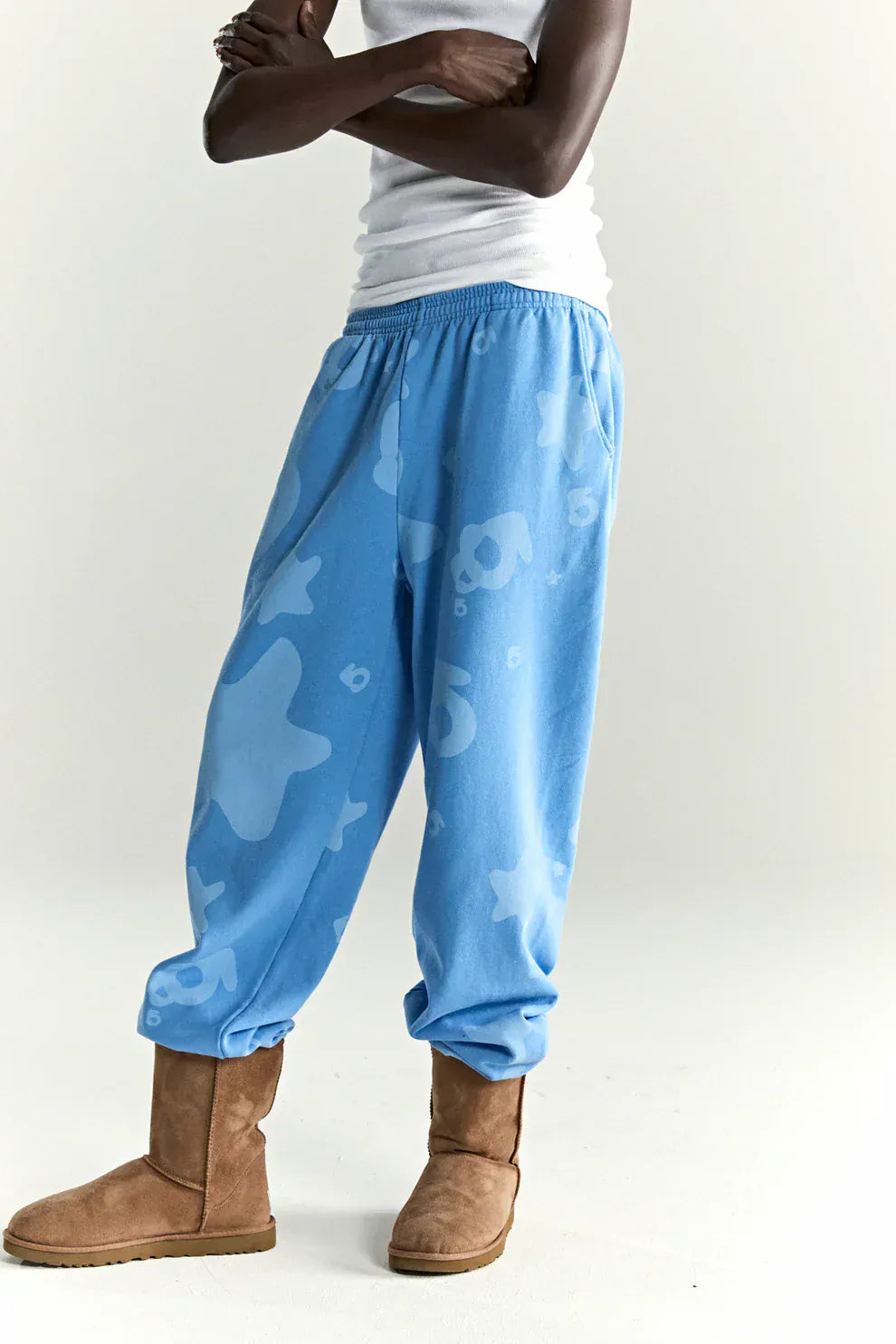 A person stands with arms crossed, wearing a white tank top, Sp5der Light Blue All-Over Print Sweatpants 100% Cotton Unisex Streetwear Joggers, and brown suede boots.