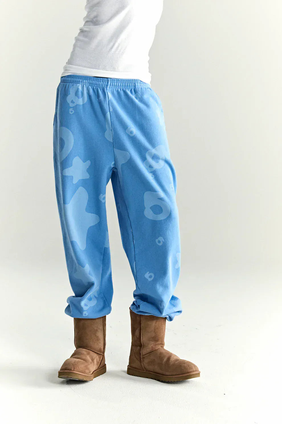 Person wearing Sp5der Light Blue All-Over Print Sweatpants—100% cotton unisex streetwear joggers—with abstract patterns and tan boots, standing against a white background. These sweatpants offer premium comfort and style.