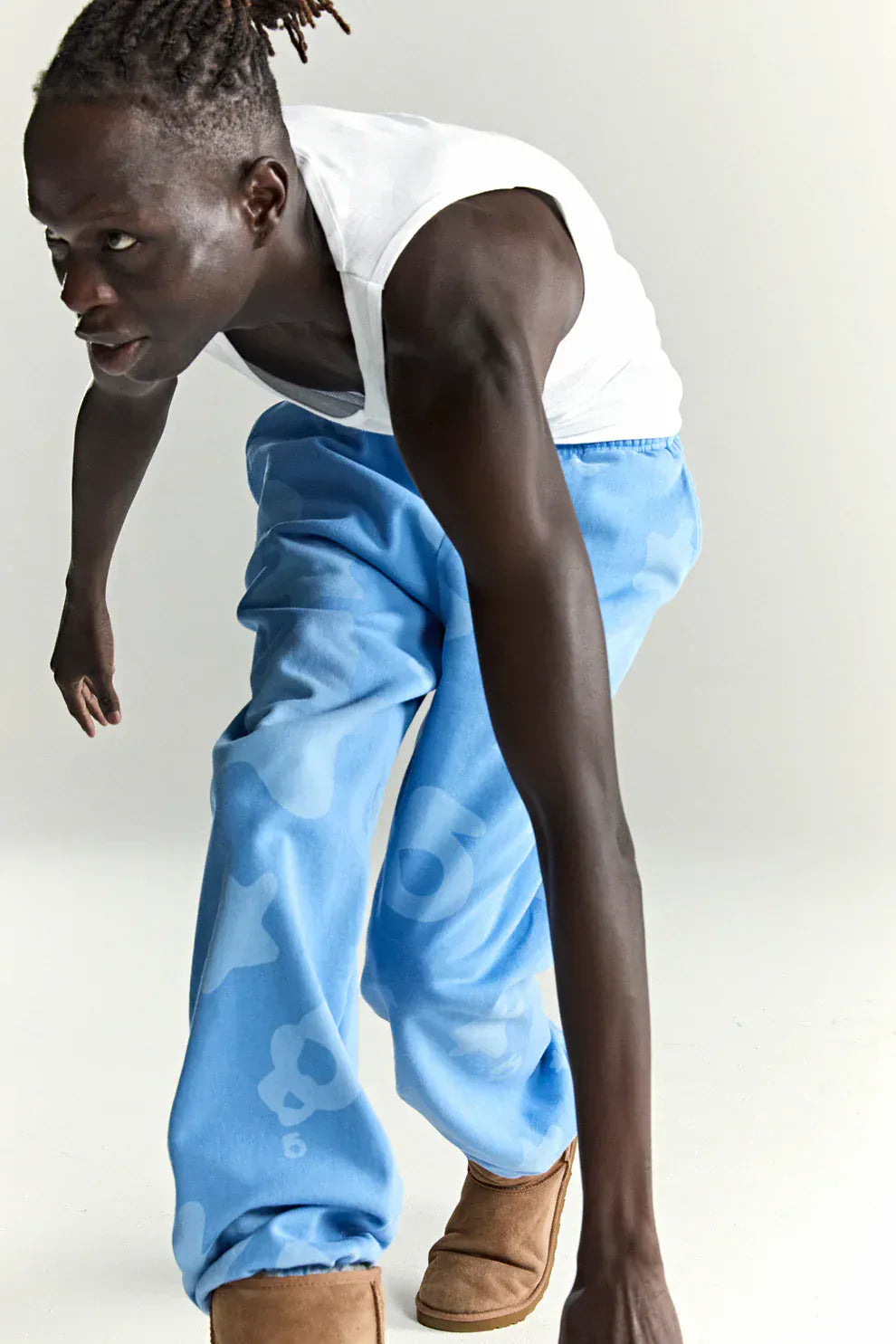 Wearing a white tank top, Sp5der Light Blue All-Over Print Sweatpants 100% Cotton Unisex Streetwear Joggers, and tan boots, a person leans forward dynamically against a plain light background.