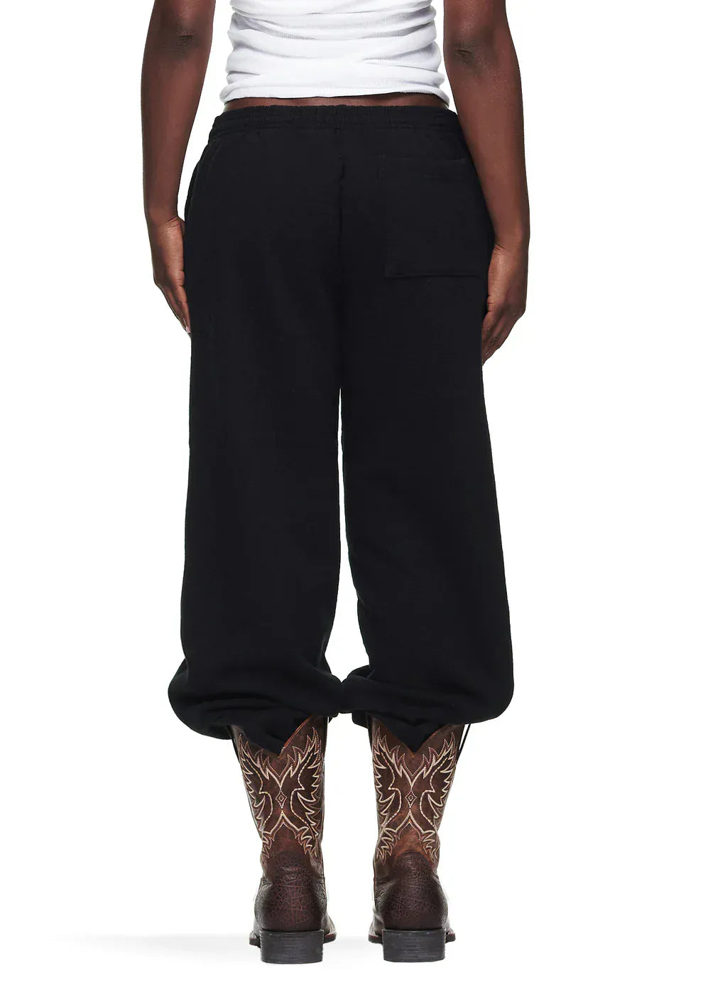 Someone stands facing away from the camera, wearing Sp5der Sweatpants – unisex black cotton sweatpants with a front logo and star print, paired with a white top and brown cowboy boots.