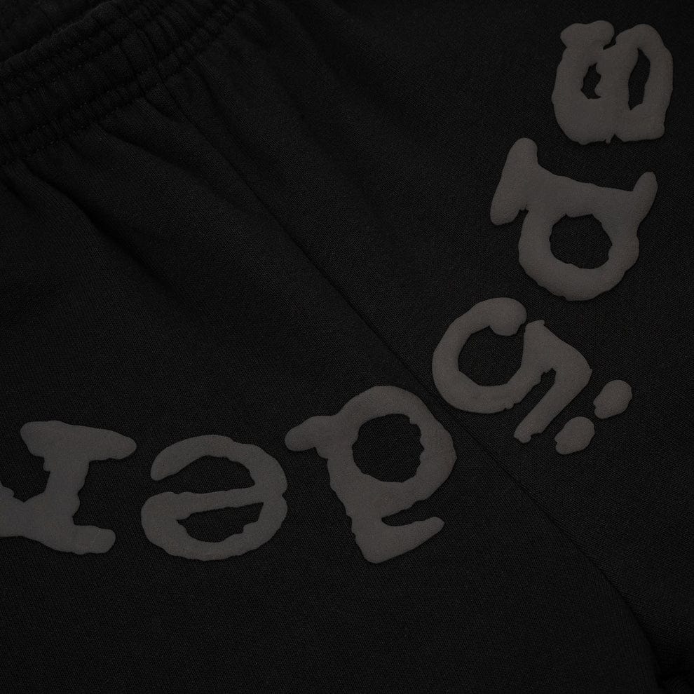 Close-up of the Sp5der Black Tonal Logo Sweatpants (100% cotton unisex streetwear joggers) shows black fabric with raised, textured gray text in a stylized font, featuring "aged" upside down—a distinctive detail.