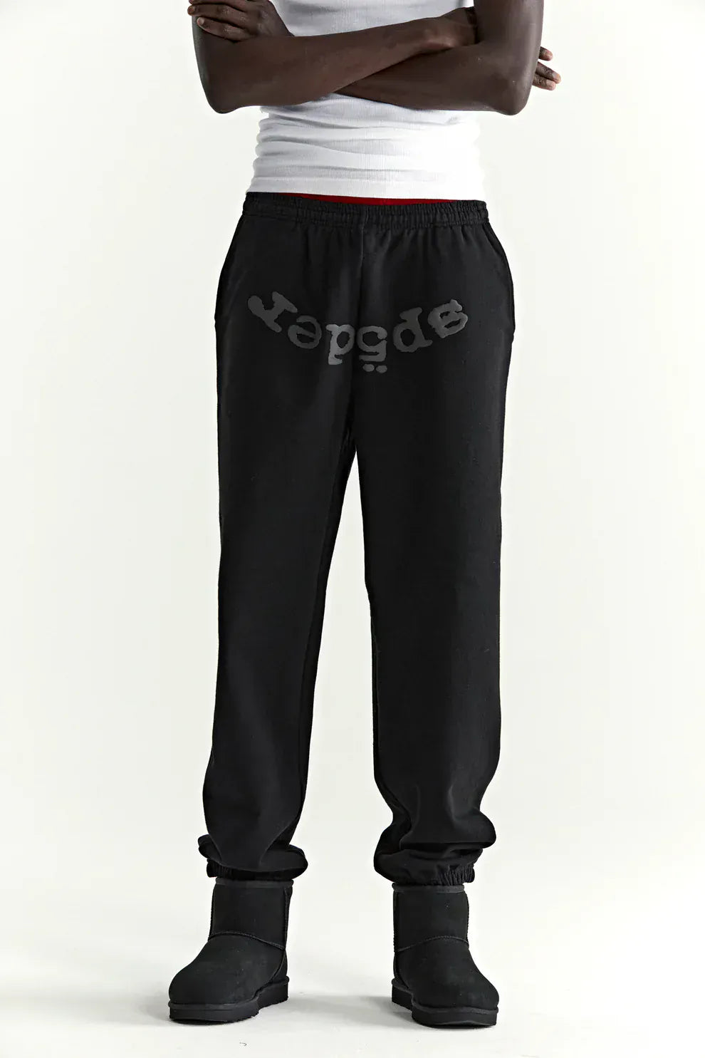 Person wearing a white tank top, Sp5der Black Tonal Logo Sweatpants 100% Cotton Unisex Streetwear Joggers, and black boots stands with arms crossed against a plain background—showcasing bold streetwear style.