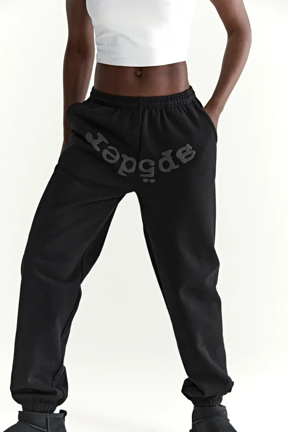 A person wears a white crop top and Sp5der Black Tonal Logo Sweatpants 100% Cotton Unisex Streetwear Joggers, with the logo printed upside down on the left leg, standing against a plain light background for an effortless streetwear look.