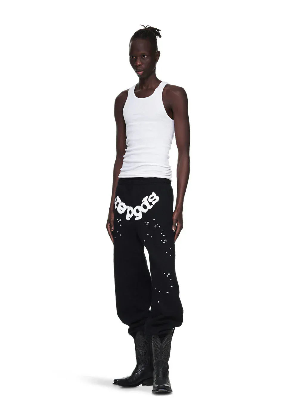A person stands facing forward in a white tank top, Sp5der Sweatpants—unisex black cotton with front logo and star print—and black cowboy boots against a plain background, creating a bold streetwear look.