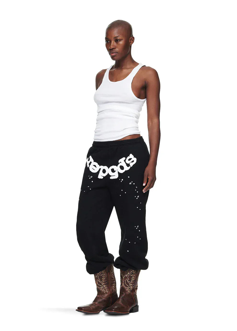 A person wearing a white tank top, Sp5der Unisex Black Cotton Sweatpants with front logo and star print, and brown cowboy boots stands against a white background.