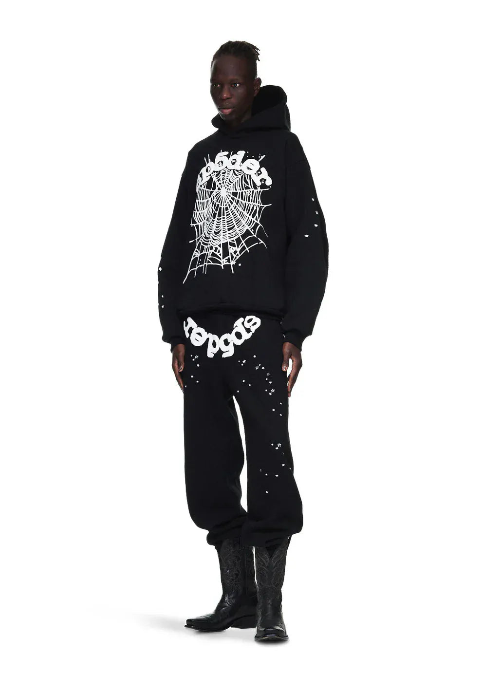 A person wears a black hoodie and Sp5der Sweatpants—unisex black cotton sweatpants with a front logo and star print—paired with black cowboy boots for a bold streetwear look, standing against a white background.