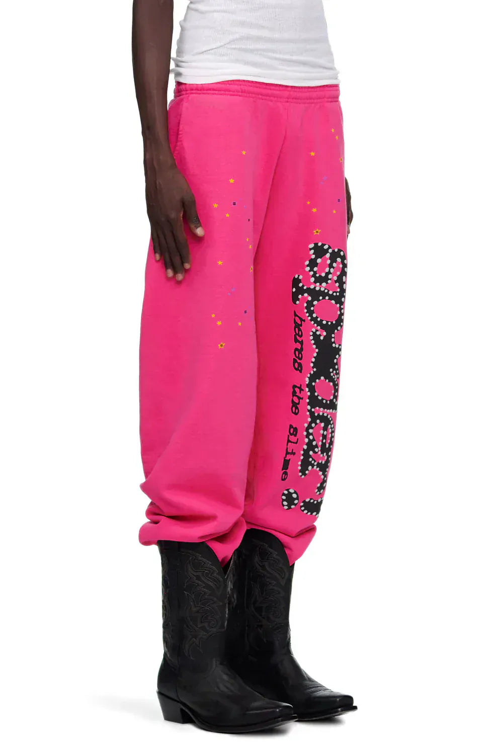 Someone stands against a plain background wearing Sp5der Sweatpants—unisex pink cotton wide leg sweatpants with a graphic logo print—paired with a white tank top and black cowboy boots.