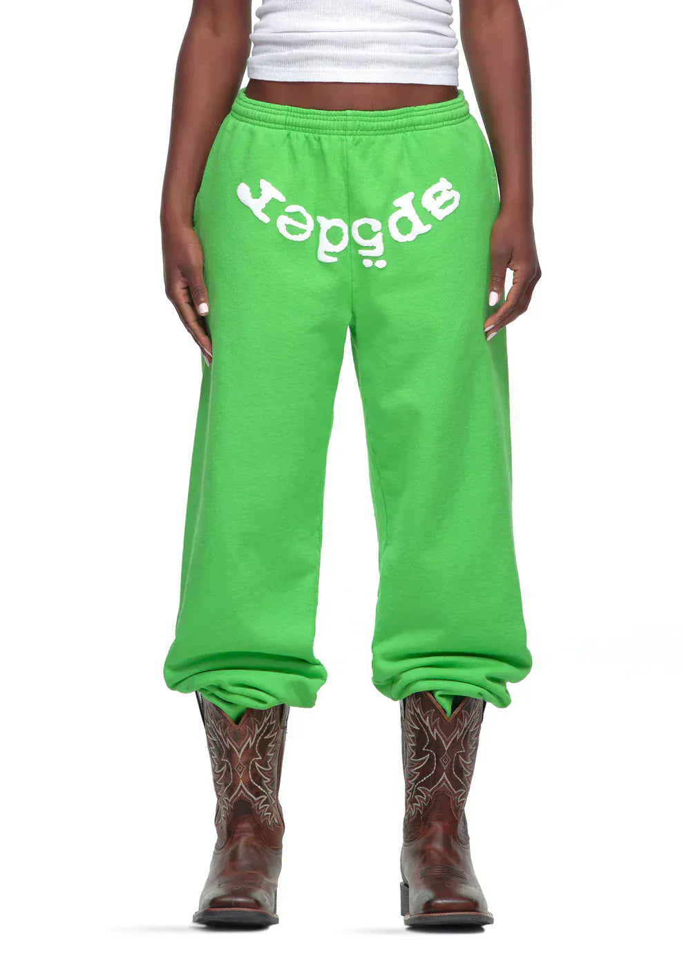 A person in Sp5der Sweatpants – Unisex Green Cotton Sweatpants with Front Logo Print, paired with a white tank top and brown cowboy boots, stands against a plain backdrop—showcasing bold streetwear style.