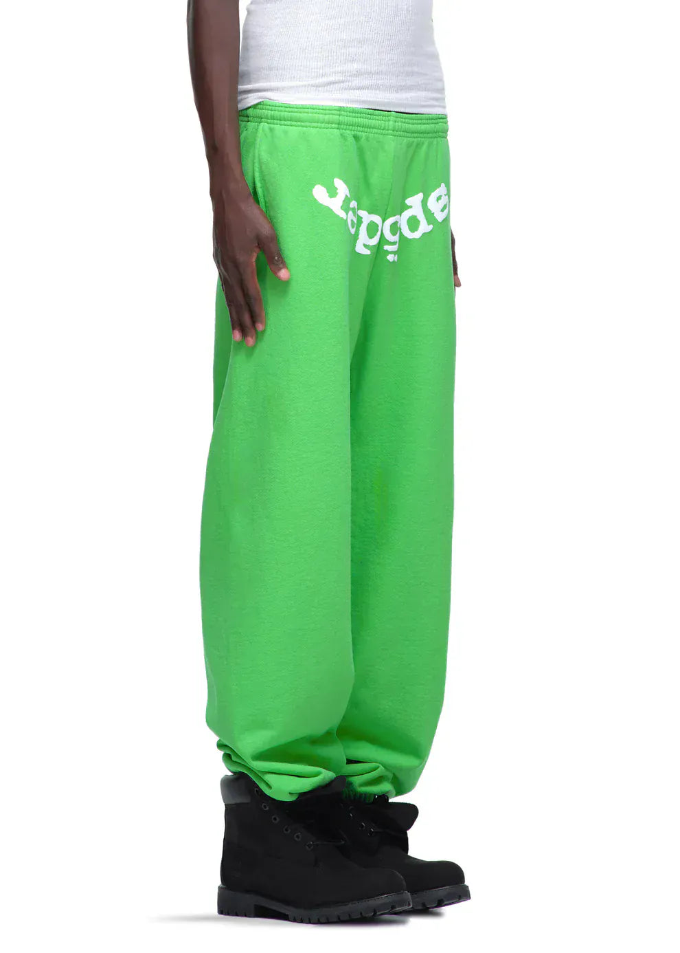 A person in Sp5der Sweatpants – Unisex Green Cotton Sweatpants with front logo print, a white tank top, and black boots, shown from the side.